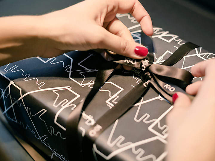 Hände mit rotem Nagellack binden eine schwarze Schleife an ein in schwarz-weißes Papier mit geometrischem Muster eingewickeltes Geschenk.