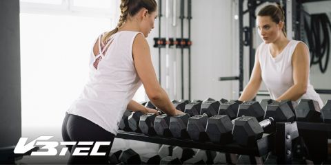 Eine Person mit geflochtenen Haaren und Trainingskleidung steht vor einem Hantelständer und betrachtet ihr Spiegelbild. In der Ecke ist das Logo „V3TEC“ zu sehen.