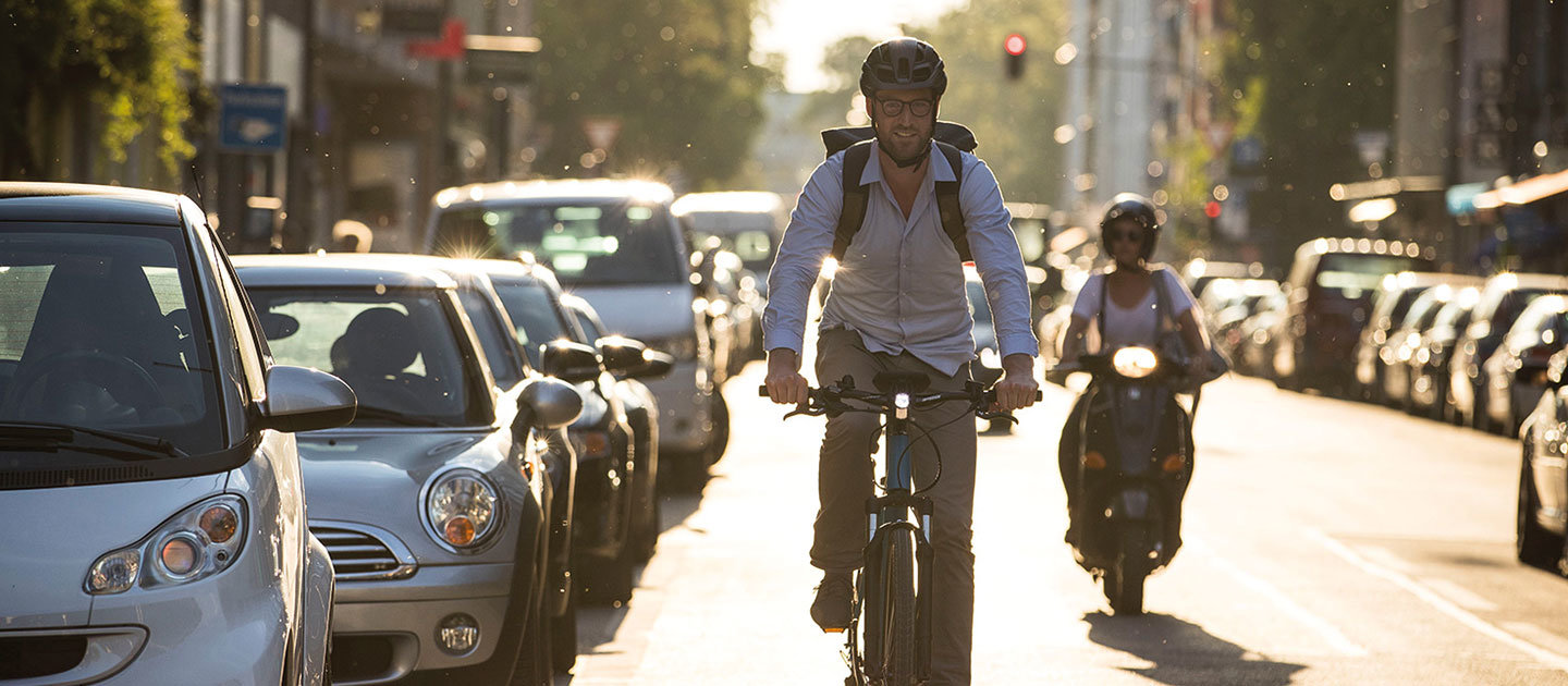 Ein Mann mit Helm und Brille fährt auf einer Stadtstraße Fahrrad, umgeben von parkenden Autos. Das Sonnenlicht erzeugt einen warmen Schein und eine Person auf einem Roller fährt hinter ihm her. Die Szene lässt auf einen städtischen Pendlerverkehr schließen.