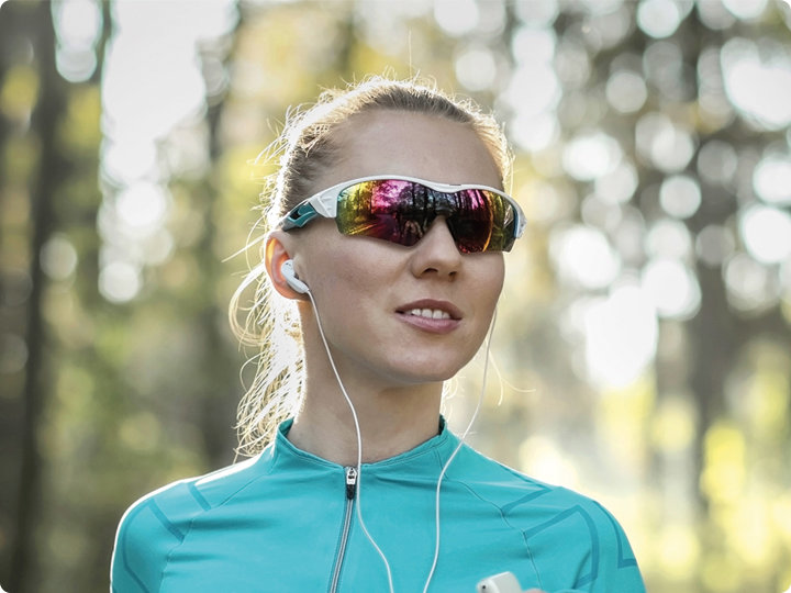Eine Person mit verspiegelter Sonnenbrille und Kopfhörern steht draußen in einem Waldstück. Sie trägt eine türkisfarbene Sportjacke und lächelt, während im Hintergrund Sonnenlicht durch die Bäume fällt.