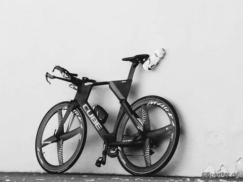 Ein elegantes, schwarzes Rennrad mit aerodynamischem Design und geschlossenen Rädern lehnt an einer schlichten Wand. Am Rahmen ist eine Wasserflasche befestigt. Das Rad trägt die Marke „Cube“ und auf den Rädern steht „Mavic“.