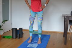 Eine Person in einem roten Hemd und gemusterten Leggings steht auf einer blauen Trainingsmatte und hat ein Widerstandsband um ihre Waden. In der Nähe befinden sich zwei Schaumstoffrollen und rechts ist teilweise ein Tisch sichtbar.