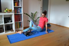 Eine Frau in bunten Leggings und einem rosa Hemd benutzt eine Schaumstoffrolle an ihrer Wade, während sie auf einer blauen Yogamatte in einem Wohnzimmer sitzt. Im Hintergrund stehen ein Bücherregal, eine Pflanze und eine Obstschale.