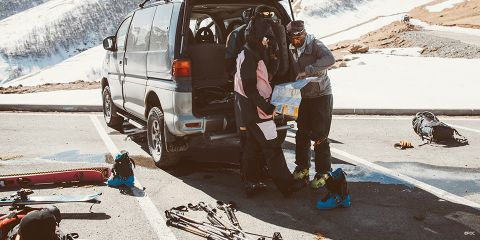 Zwei Personen in Skiausrüstung studieren auf einem verschneiten Parkplatz neben ihrem Van eine Karte. Skier und Stöcke liegen auf dem Boden, die umgebende Landschaft ist verschneit. Um sie herum sind Skier, Stiefel und Rucksäcke zu sehen.
