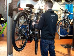 Ein Mechaniker in einer Werkstatt justiert ein schwarzes Elektrofahrrad, das auf einem Ständer montiert ist. Der Raum ist mit Werkzeugen gefüllt und im Hintergrund ist ein weiteres Fahrrad zu sehen. Der Mechaniker trägt Jeans und eine dunkle Jacke mit der Nummer „1“ auf dem Rücken.