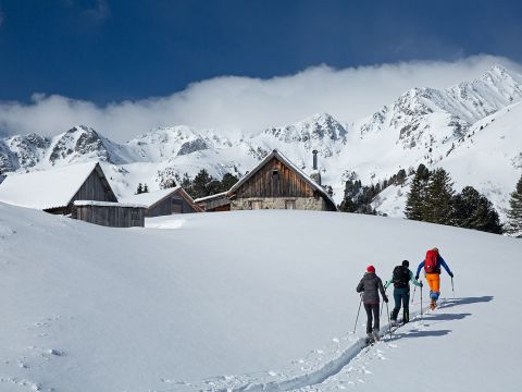 Drei Menschen mit Rucksäcken und Skistöcken wandern durch eine verschneite Landschaft zu urigen Hütten. Im Hintergrund sind schneebedeckte Berge und ein klarer, tiefblauer Himmel zu sehen.