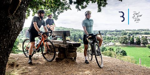 Drei Radfahrer mit Helmen und lässiger Radsportkleidung stehen neben einer Holzbank auf einem Hügel und blicken auf eine grüne Landschaft und ein Dorf. Über ihnen spendet ein Baum Schatten. Der Himmel ist teilweise bewölkt. Auf der Seite steht: „Teil Ihrer Fahrt.“.