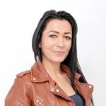 Frau mit langen dunklen Haaren, trägt eine braune Lederjacke und ein dunkles Oberteil und lächelt leicht vor einem schlichten weißen Hintergrund.