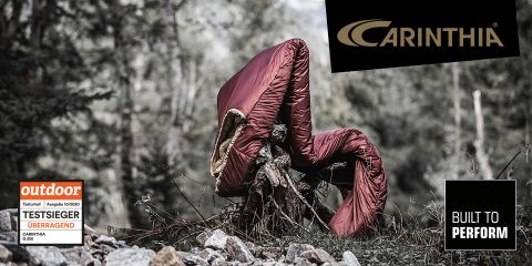 Ein kastanienbrauner Schlafsack hängt über einem Baumstumpf in einer Waldlandschaft. Oben rechts ist das Carinthia-Logo zu sehen. Außerdem sind ein Testsieger-Sieger der Zeitschrift „Outdoor“ und der Schriftzug „Built to Perform“ zu sehen.
