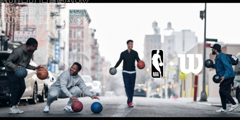 Vier Leute spielen Basketball auf einer Stadtstraße, jeder dribbelt einen Ball. Die Straße ist von Gebäuden gesäumt und der Himmel ist bedeckt. In der Mitte sind die Logos von NBA und Wilson zu sehen. Oben stehen die Worte „BUILD LEGACY“.