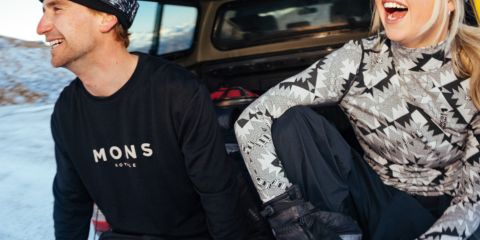 Zwei Personen in Winterkleidung sitzen hinten in einem auf einer verschneiten Straße geparkten Fahrzeug. Beide lächeln. Die Person links trägt eine schwarze Mütze und ein Sweatshirt, während die Person rechts einen gemusterten Pullover, einen Helm und eine Schutzbrille trägt. Text auf dem Bild.