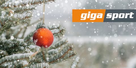 Im Vordergrund hängt ein schneebedeckter immergrüner Zweig mit einem roten Weihnachtsornament. Leise fällt Schnee und in der oberen rechten Ecke steht auf einem grau-orangefarbenen Hintergrund „Giga Sport“.
