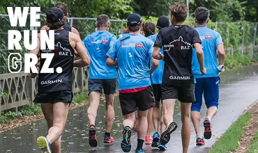 Eine Gruppe Läufer auf einem Parkweg. Einige tragen blaue Shirts, andere schwarze mit der Aufschrift „RUNNING RAZ“. Bäume und ein Zaun säumen den Weg. In der oberen linken Ecke steht in Weiß „WE RUN GRZ.“.