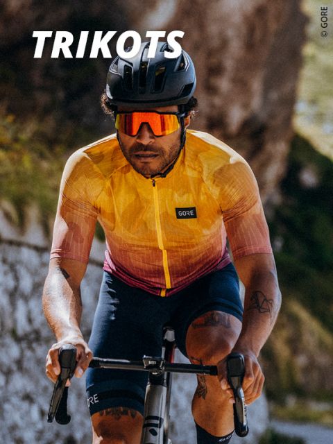 Radfahrerin in einem gelb-roten Trikot mit „GORE“-Schriftzug fährt im Freien. Sie hat dunkles, lockiges Haar, trägt eine Sonnenbrille und einen Helm. Sonnenlicht hebt die Tattoos an den Armen hervor. Im Hintergrund ist eine verschwommene Naturlandschaft zu sehen. Der Text lautet „TRIKOTS“.