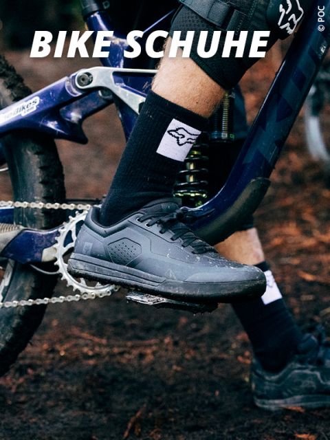 Eine Person mit schwarzen Radschuhen und Socken fährt auf einem blauen Mountainbike. Der Boden ist mit Erde bedeckt, oben drauf steht „BIKE SCHUHE“.