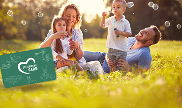 Eine vierköpfige Familie genießt einen sonnigen Tag auf einer Wiese. Zwei Kinder machen Seifenblasen, während die Eltern lächelnd zuschauen. In der unteren linken Ecke befindet sich eine grüne Karte mit weißen Symbolen von Sehenswürdigkeiten.