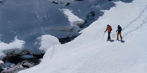 Zwei Personen wandern durch eine verschneite Landschaft. Sie tragen Winterkleidung und Rucksäcke sowie Schneeschuhe oder Skier. Ein Bach ist teilweise unter dem Schnee sichtbar, und in der Nähe sind Tierspuren zu sehen. Das Sonnenlicht wirft hohe Schatten.