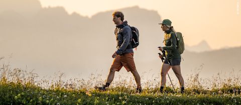Zwei Menschen mit Rucksäcken und Outdoor-Kleidung wandern auf einem grasbewachsenen Pfad mit Wildblumen vor der Kulisse von Bergen in sanftem, goldenem Licht.