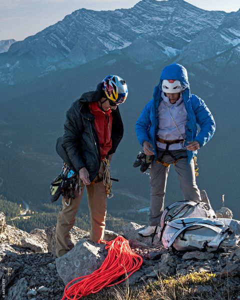 Zwei Bergsteiger mit Helmen und Jacken stehen an einem felsigen Berghang und holen Ausrüstung und Seile aus einem Rucksack, im Hintergrund eine malerische Berglandschaft.