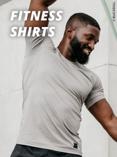 Ein lächelnder Mann, der ein hellgraues Fitness-Shirt trägt, streckt seinen Arm über seinen Kopf. Der Text "FITNESS SHIRTS" ist in fetten weißen Buchstaben auf dem Bild zu sehen.