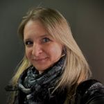 Eine Frau mit langen blonden Haaren lächelt leicht in die Kamera. Sie trägt einen schwarz gemusterten Schal und eine schwarze Jacke vor einem dunklen, schlichten Hintergrund.