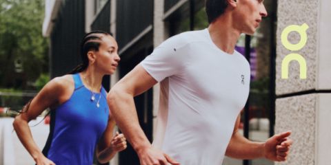 Zwei Personen in Sportkleidung laufen im Freien neben einem Gebäude. Der Vordermann trägt ein weißes Hemd, der Hintermann ein blaues Oberteil. Das Logo der Sportmarke "On" erscheint auf der rechten Seite des Bildes.