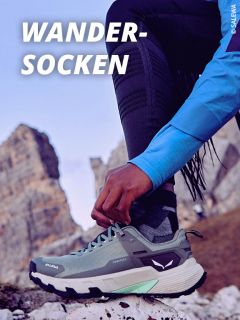 Eine Person mit Trailrunning-Schuhen und gemusterten Outdoor-Socken passt ihr Schuhwerk auf einem felsigen Berggelände an, mit zerklüfteten Gipfeln im Hintergrund. Der Text "OUTDOOR-SOCKEN" erscheint oben links.