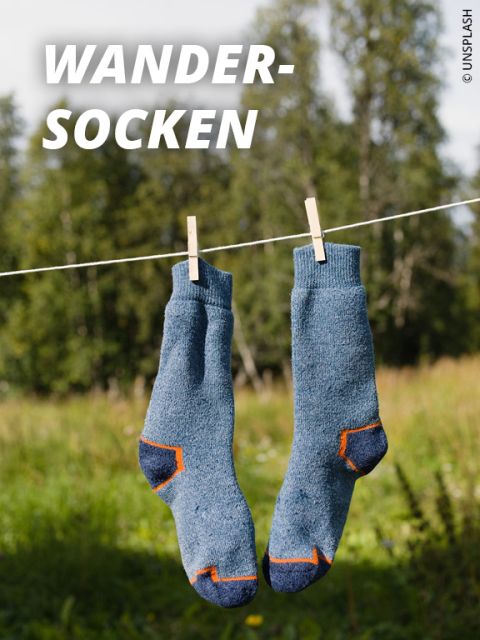 Ein Paar blaue Outdoor-Socken mit orangefarbenen Akzenten hängen an einer Wäscheleine mit Wäscheklammern. Der Hintergrund zeigt grünes Gras, Bäume und einen sonnigen Himmel. Der Text lautet "OUTDOOR-SOCKEN.