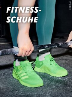 Person in hellgrünen Adidas-Turnschuhen, die eine Hantel hält und bereit ist, Gewichte zu stemmen. Der Text auf Deutsch lautet "Fitness-Schuhe". Beine und Füße sind sichtbar, was auf ein Fitnessstudio oder eine Trainingsumgebung hindeutet.
