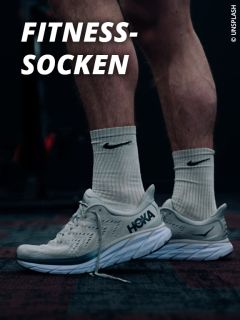 Eine Person mit weißen Nike-Socken und hellgrauen Hoka-Laufschuhen steht auf dem Boden eines dunklen Fitnessstudios. Oben links steht in fetter weißer Schrift "FITNESS-SOCKEN".