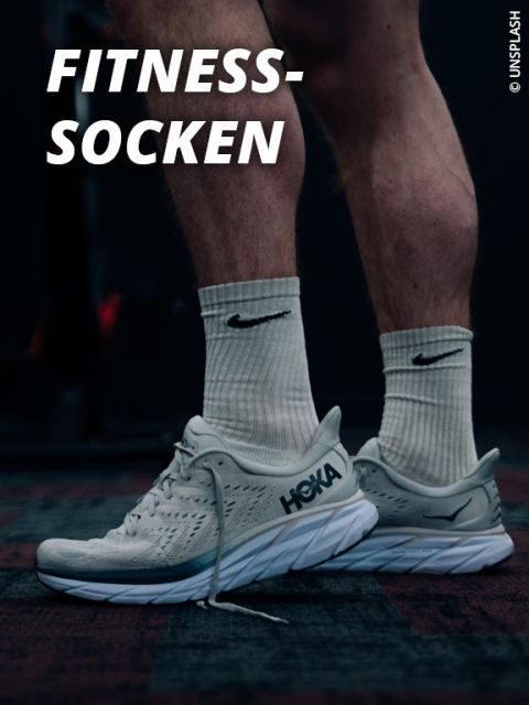 Eine Person mit weißen Nike-Socken und hellgrauen Hoka-Laufschuhen steht auf dem Boden eines dunklen Fitnessstudios. Oben links steht in fetter weißer Schrift "FITNESS-SOCKEN".