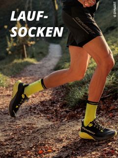 Eine Person, die auf einem Waldweg läuft, trägt gelbe Dynafit-Laufsocken und schwarze Laufschuhe. Der Text "LAUF-SOCKEN" erscheint in fetten weißen Buchstaben in der oberen linken Ecke.