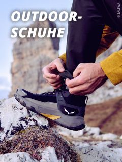 Person, die die Schnürsenkel eines schwarzen Outdoor-Wanderschuhs auf einer felsigen Oberfläche bindet, wobei im unscharfen Hintergrund Berge zu sehen sind. Der Text "OUTDOOR-SCHUHE" erscheint in fetten weißen Buchstaben oben links.