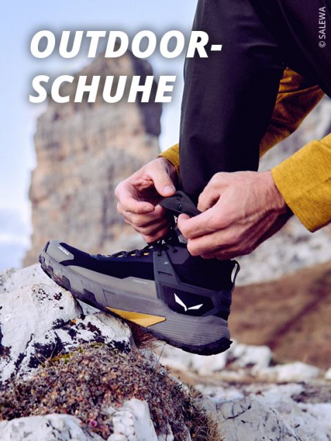Person, die die Schnürsenkel eines schwarzen Outdoor-Wanderschuhs auf einer felsigen Oberfläche bindet, wobei im unscharfen Hintergrund Berge zu sehen sind. Der Text "OUTDOOR-SCHUHE" erscheint in fetten weißen Buchstaben oben links.