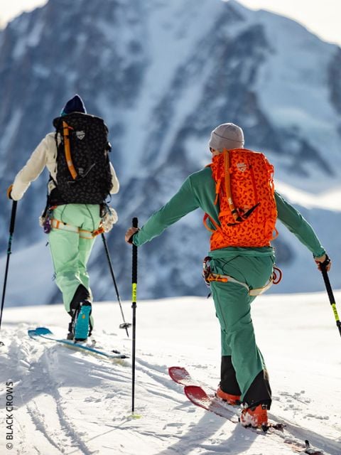 Zwei Personen in bunten Skiklamotten und Rucksäcken fahren auf einem verschneiten Berg bergauf und stützen sich dabei mit Stöcken ab. Im Hintergrund ist ein großer, schroffer Berggipfel zu sehen.