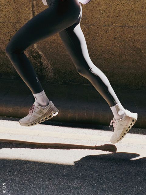 Eine Person, die hellbeige Sportschuhe mit weißen Sohlen und eine schwarze Hose trägt, wird beim Springen mit den Füßen auf dem Boden vor einem dunklen Hintergrund aufgenommen.