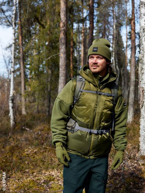 Ein Mann mit einer olivgrünen Mütze, Jacke und Handschuhen steht in einem sonnenbeschienenen Wald und schaut zur Seite. Er trägt einen Rucksack und ist von hohen Bäumen und spärlichem Gestrüpp umgeben.