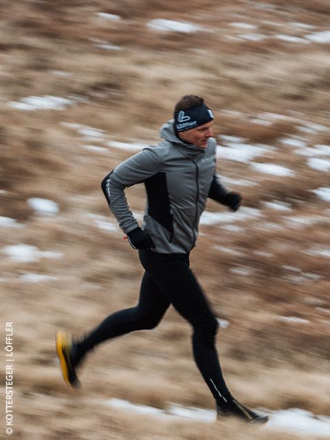 Eine Person in Sportkleidung läuft im Freien auf einem verschwommenen, grasbewachsenen Weg mit Schneeflecken, bekleidet mit einer grauen Jacke, schwarzen Leggings und einem Stirnband.