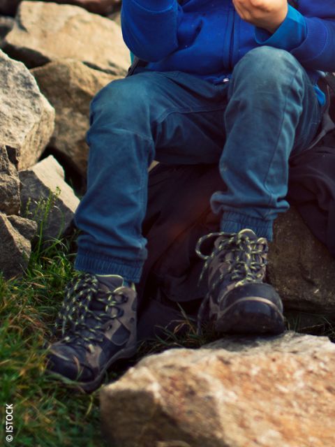 Zwei Jungen sitzen im Freien auf einem Felsen. Ein Junge in einer blauen Jacke lächelt, während er einen Snack isst, und der andere, in einer grünen Jacke, schaut nach unten und sucht in seinem Rucksack. Beide tragen Wanderschuhe und Rucksäcke.