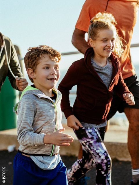 Zwei lächelnde Kinder, ein Junge und ein Mädchen, joggen in sportlicher Kleidung im Freien. Erwachsene laufen neben ihnen her. Die Szene ist sonnig und energiegeladen und deutet auf einen Familien- oder Gruppenlauf hin.
