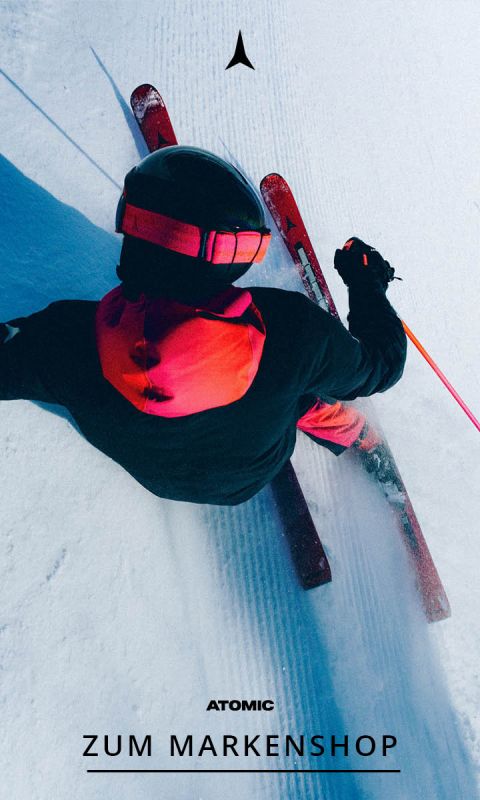Ein Skifahrer in einem schwarz-roten Outfit gleitet einen verschneiten Hang hinunter und hinterlässt dabei Spuren im Schnee. Die Ansicht ist von oben, und der Text am unteren Rand lautet "Atomic Zum Markenshop".