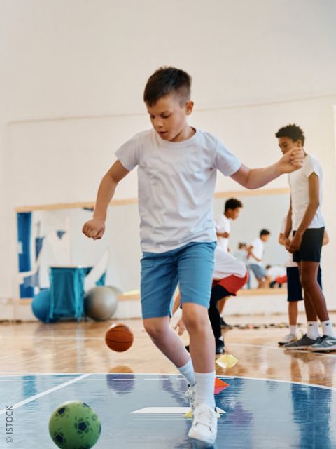 Ein Junge in einem weißen T-Shirt und blauen Shorts balanciert auf einem Fuß, während er in einer Turnhalle spielt. Im Hintergrund sind andere Kinder und Sportbälle zu sehen.