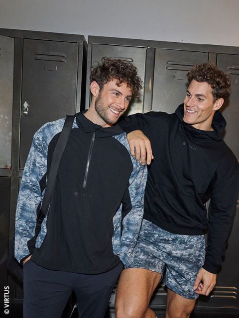 Zwei junge Männer in Sportkleidung lächeln und posieren gemeinsam vor einem Spind. Der eine trägt eine schwarz-blaue Jacke und eine Umhängetasche, der andere einen schwarzen Kapuzenpulli und gemusterte Shorts und stützt seinen Arm auf die Schulter seines Freundes.