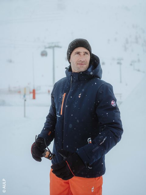Ein Mann in einer marineblauen Winterjacke, einer orangefarbenen Hose, einer schwarzen Mütze und Handschuhen steht in einer verschneiten Umgebung im Freien und hält eine Sonnenbrille. Im Hintergrund sind Skilifte und schneebedeckte Pisten zu sehen.