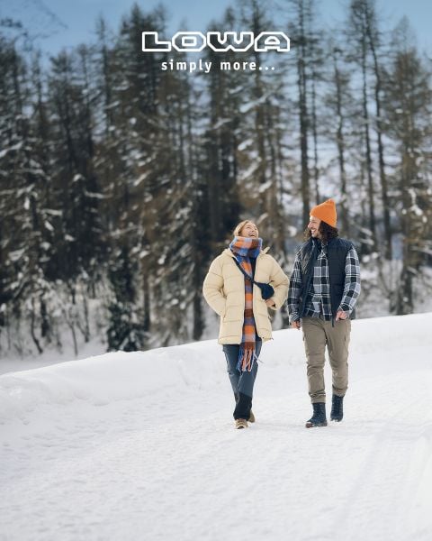 Eine Frau und ein Mann gehen gemeinsam auf einem verschneiten Weg, in Winterkleidung und lächelnd. Schneebedeckte Bäume säumen den Hintergrund. Über ihnen erscheint der Text "LOWA einfach mehr...".