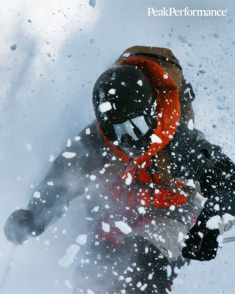 Eine Person springt unter strahlend blauem Himmel zwischen großen Felsen in einer Bergregion. Sie trägt Sportkleidung, darunter eine Jacke und Shorts. Oben ist der Schriftzug „Peak Performance“ zu sehen.