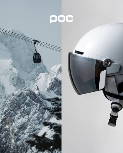 Eine Person in orangefarbenem Wintermantel und Helm steht auf einem schneebedeckten Berggipfel und blickt auf die fernen schneebedeckten Berge. Das POC-Logo ist auf dem Helm und in der oberen rechten Ecke des Bildes zu sehen.