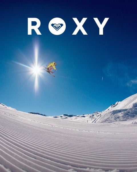 Ein Skifahrer in gelber Ausrüstung springt bei strahlend blauem Himmel und heller Sonne über eine verschneite, präparierte Bergpiste. Der Schriftzug "ROXY" mit dem Logo des Unternehmens ist oben deutlich zu sehen.