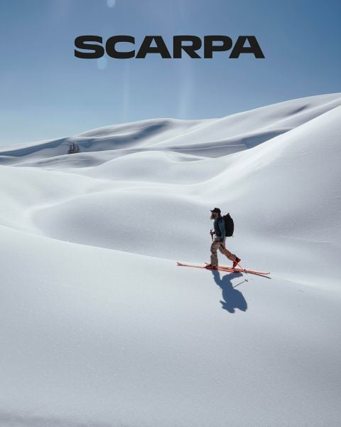 Eine Frau und ein Mann sitzen im Freien auf großen Felsen. Sie tragen Freizeitkleidung und Sonnenbrillen. Sie sind von Grün und Felsen umgeben, der Himmel ist leicht bewölkt. Oben im Bild ist das Wort „SCARPA“ zu sehen.