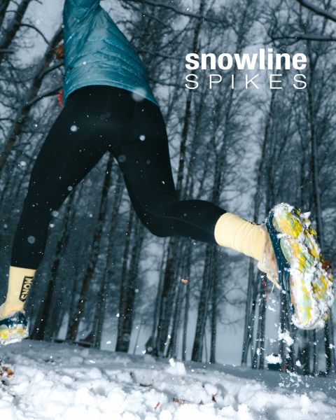 Eine Person, die Schneespikes an ihren Schuhen trägt, läuft durch einen verschneiten Wald mit hohen, blattlosen Bäumen. Der Text "snowline SPIKES" erscheint in der oberen rechten Ecke. Schnee fällt und der Boden ist weiß bedeckt.